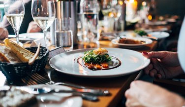 Im Restaurant steht ein Teller mit einem schön angerichtetem Fischgericht auf dem Tisch. Im Hintergrund stehen Weingläser.